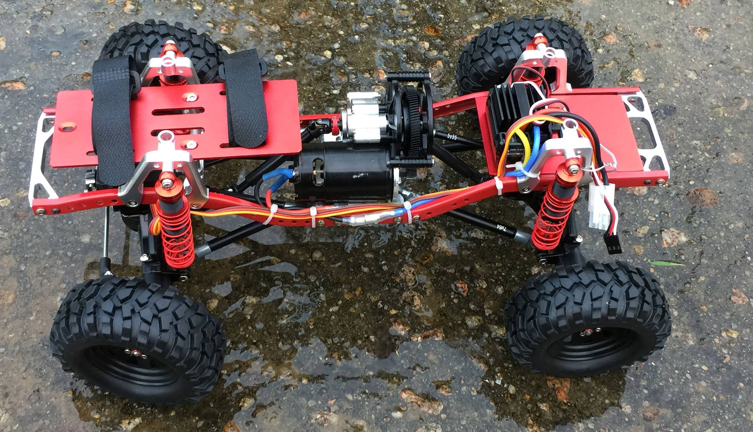 Voiture télécommandée sans carrosserie, montrant le moteur, les amortisseurs et le câblage. Le véhicule est posé sur un sol humide et pierreux.