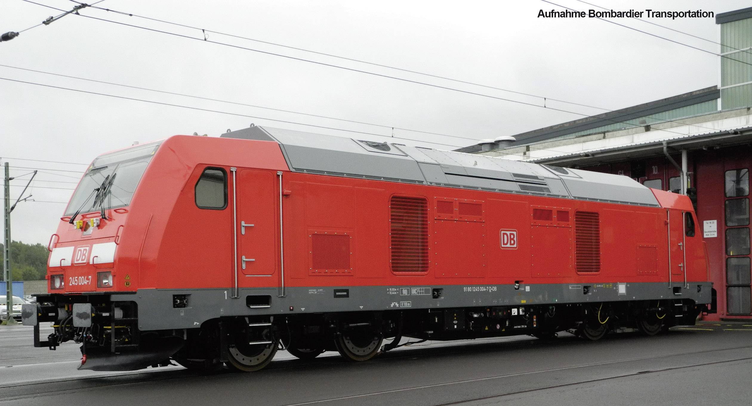 Un grand train rouge de la Deutsche Bahn est stationné sur le terrain ferroviaire à côté d'un bâtiment. Le ciel est nuageux.