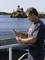 Un homme se tient sur un bateau et examine un objet dans ses mains. Un phare est visible en arrière-plan, sur une petite île.
