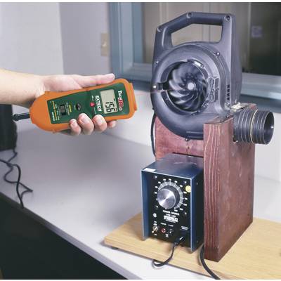 Une personne mesure le débit d'air d'un ventilateur avec un anémomètre dans un laboratoire. L'anémomètre indique '7.56'.