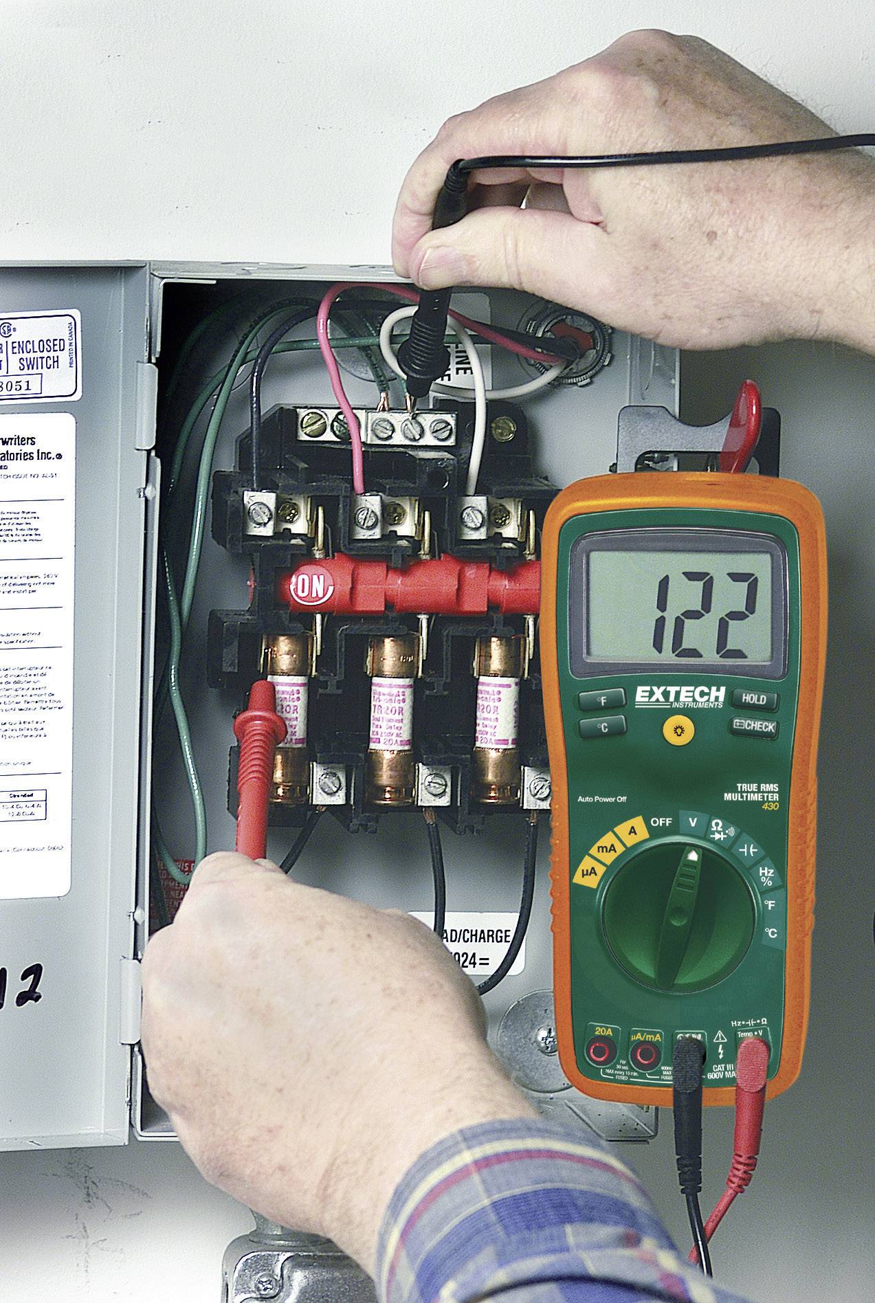Un électricien mesure la tension à l'aide d'un multimètre dans un boîtier de fusibles ouvert. L'affichage indique '122'.