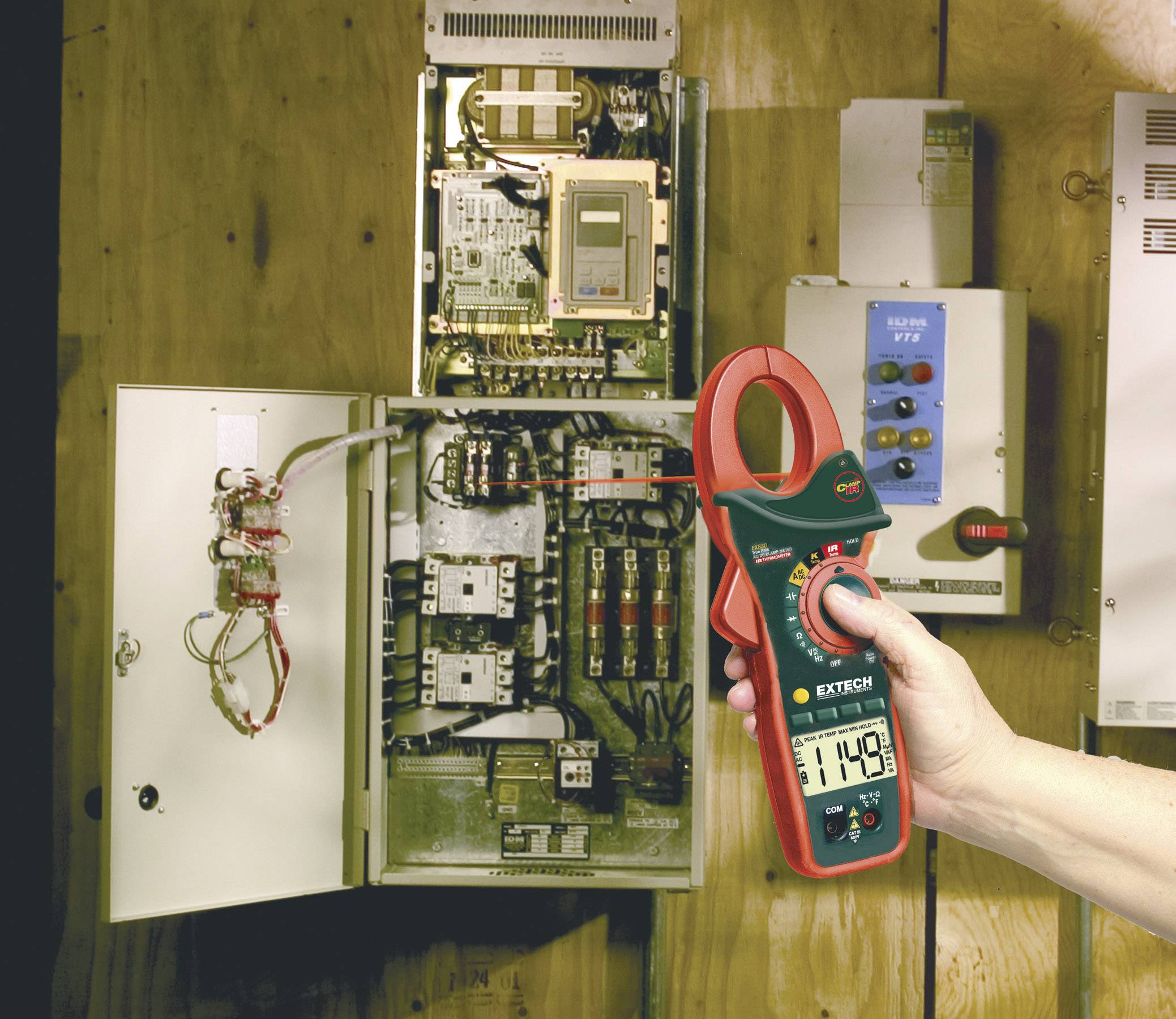 Une main tient un multimètre numérique affichant une tension de 114,9 V devant un boîtier de distribution électrique ouvert.