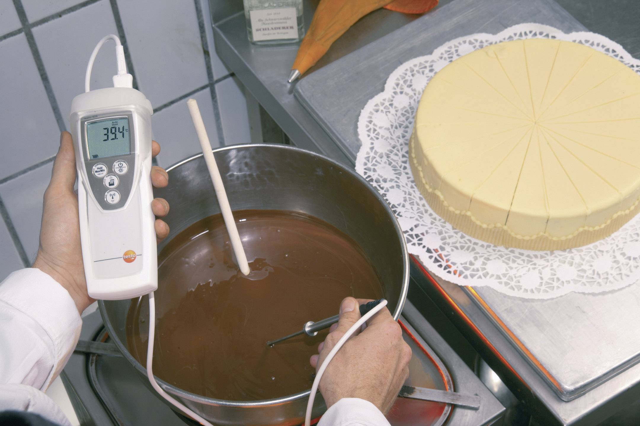 Une personne mesure la température du chocolat liquide dans une casserole à l'aide d'un thermomètre. Un gâteau clair est posé sur une table.