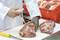 Personne en blouse blanche vaporisant un liquide sur de la viande crue sur une planche à découper. À l'arrière-plan, des morceaux de viande dans une boîte rouge.
