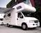Un camping-car est stationné sur un parking pavé avec son auvent déployé. Un bâtiment moderne est visible en arrière-plan.