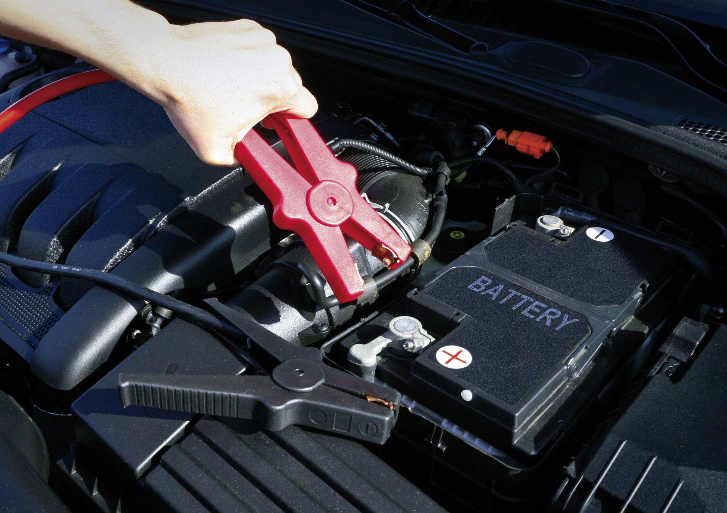 Une main connecte des câbles de démarrage rouges à une batterie de voiture. Le câble est branché sur la borne positive.
