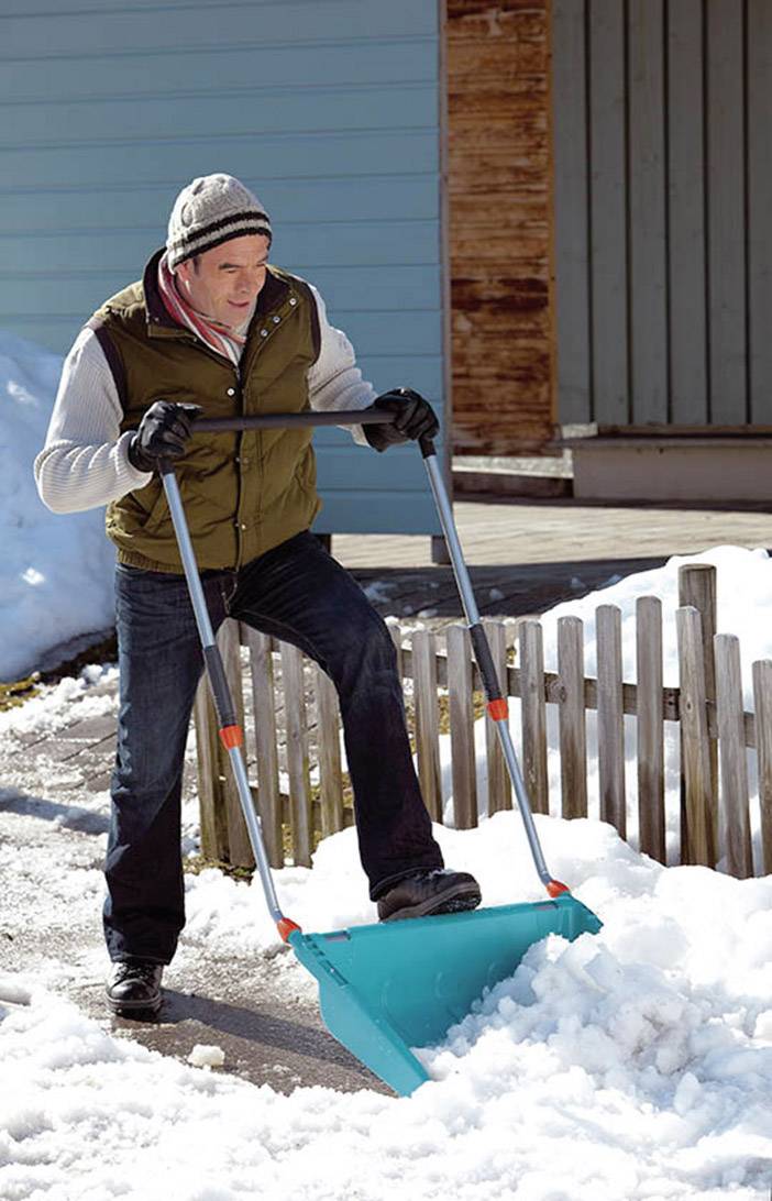 Un homme dégage la neige avec une pelle à neige sur un trottoir devant une maison. Il porte une veste d'hiver, un bonnet et des gants.