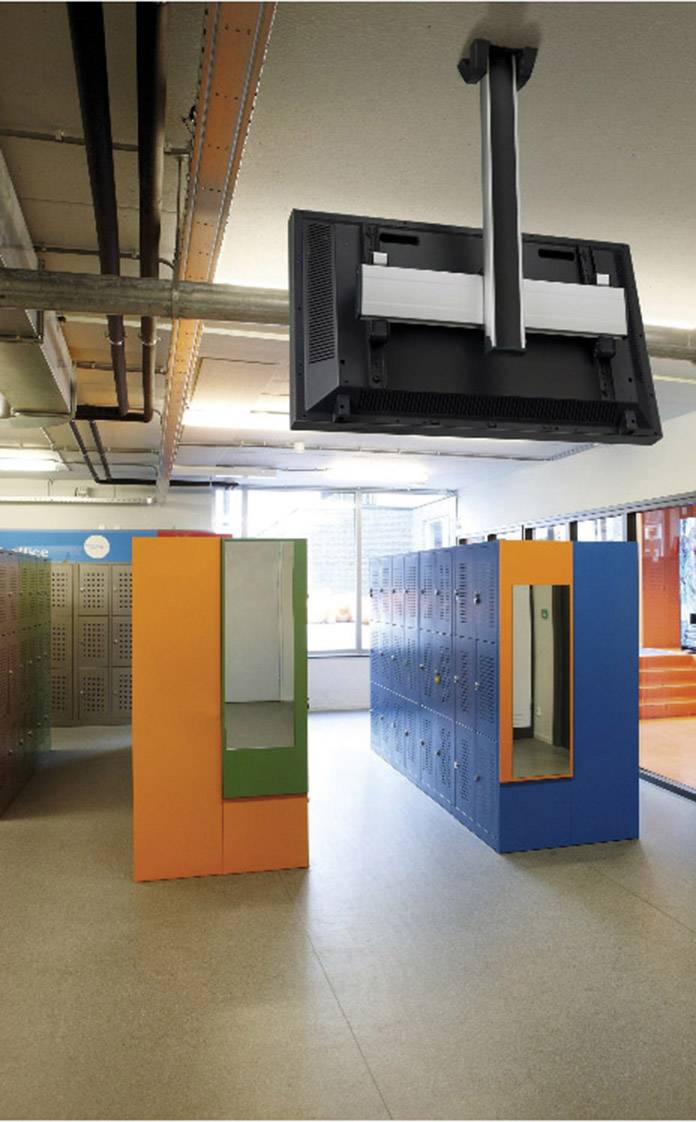 Vestiaire avec des casiers colorés. Un écran est monté au plafond. Des fenêtres à l'arrière-plan laissent entrer la lumière du jour.