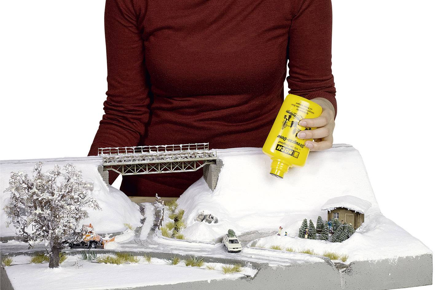 Une personne tient une bouteille jaune et applique un liquide sur un modèle de paysage hivernal avec une route, un pont et des maisons.