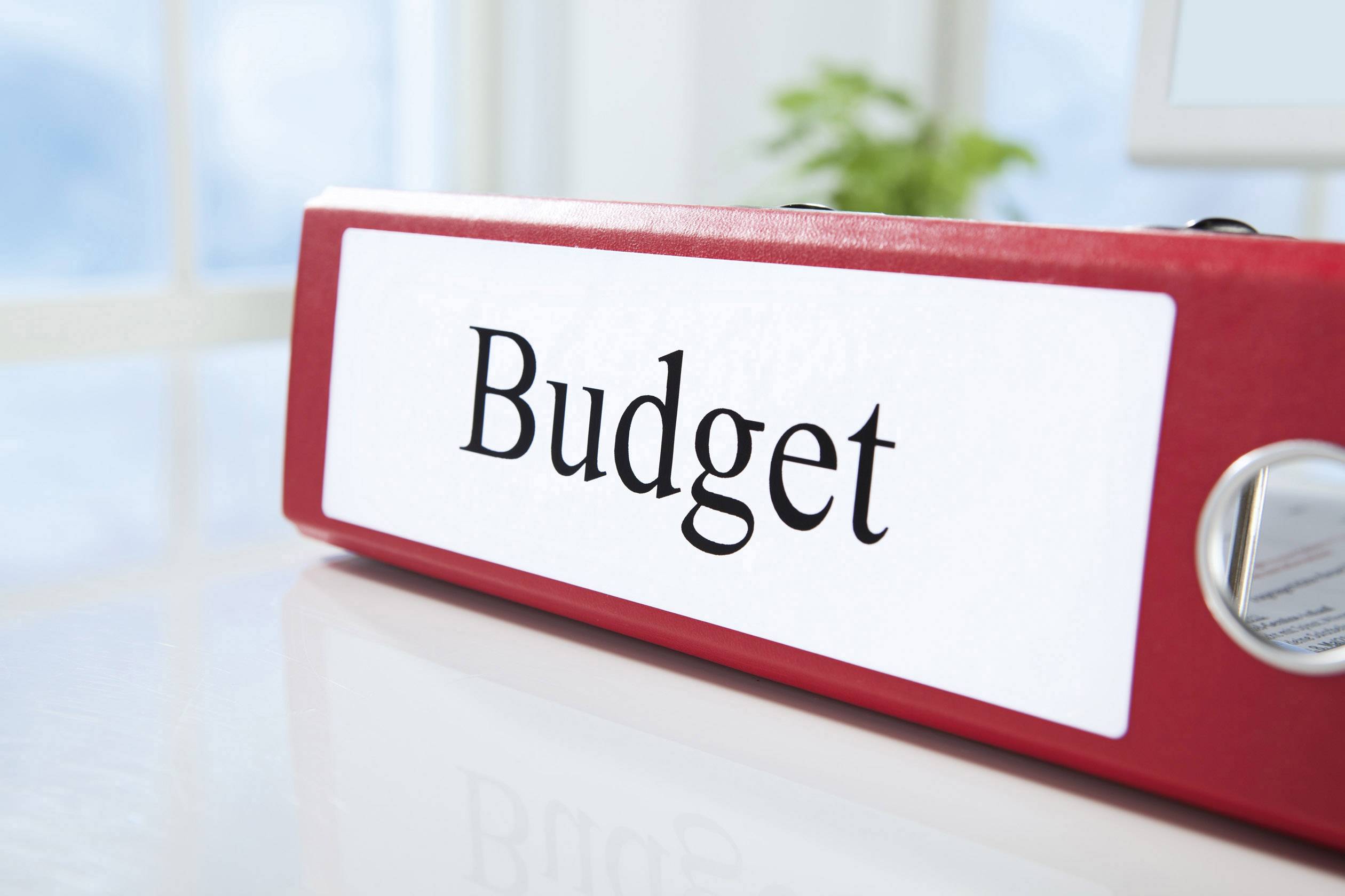 Classeur rouge portant l'inscription 'Budget' sur un bureau de bureau, avec en arrière-plan flou une fenêtre et des plantes.