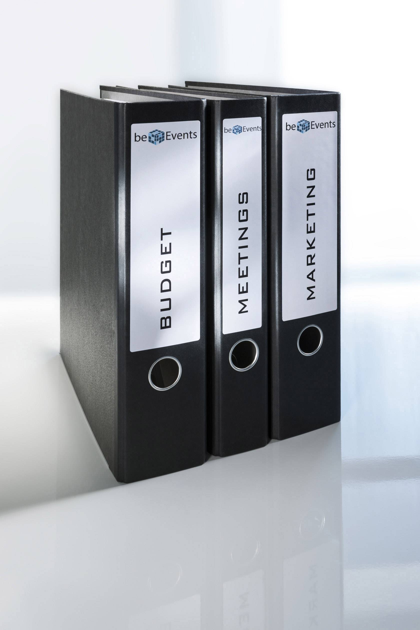 Trois classeurs noirs portant les étiquettes « Budget », « Réunions » et « Marketing » sont alignés côte à côte sur une table blanche.