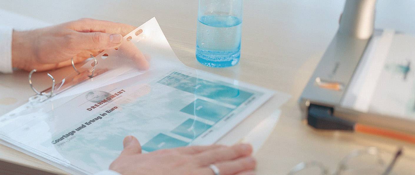 Une personne feuillette un document dans une pochette transparente posée sur un bureau, à côté d'un ordinateur portable et d'un verre d'eau.