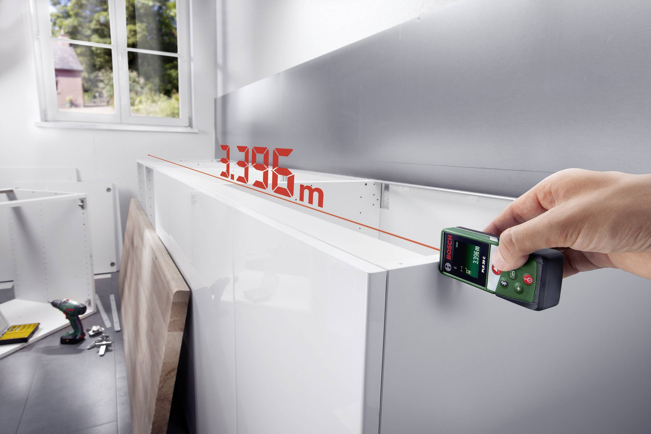 Une main tient un télémètre laser qui mesure 3,395 m et pointe vers un plan de travail de cuisine. Des outils et des matériaux sont visibles en arrière-plan.