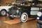 Une voiture ancienne, vert foncé, est exposée dans un hall sur un plancher en bois. À côté du véhicule, d'autres voitures classiques sont visibles.