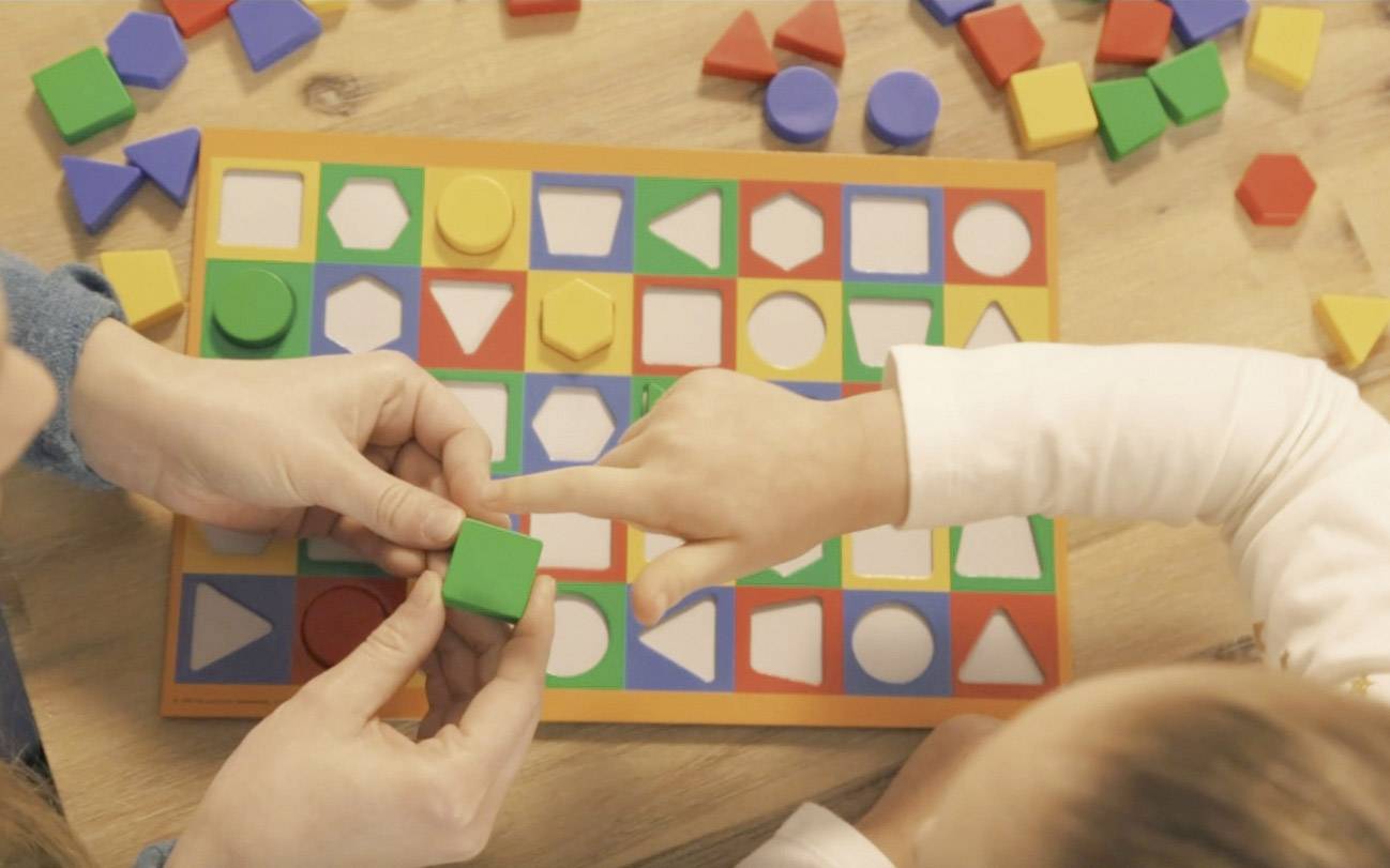 Un enfant joue avec un puzzle à encastrement coloré, disposant des formes colorées sur un modèle adapté. Les mains d'un adulte aident à l'assemblage.