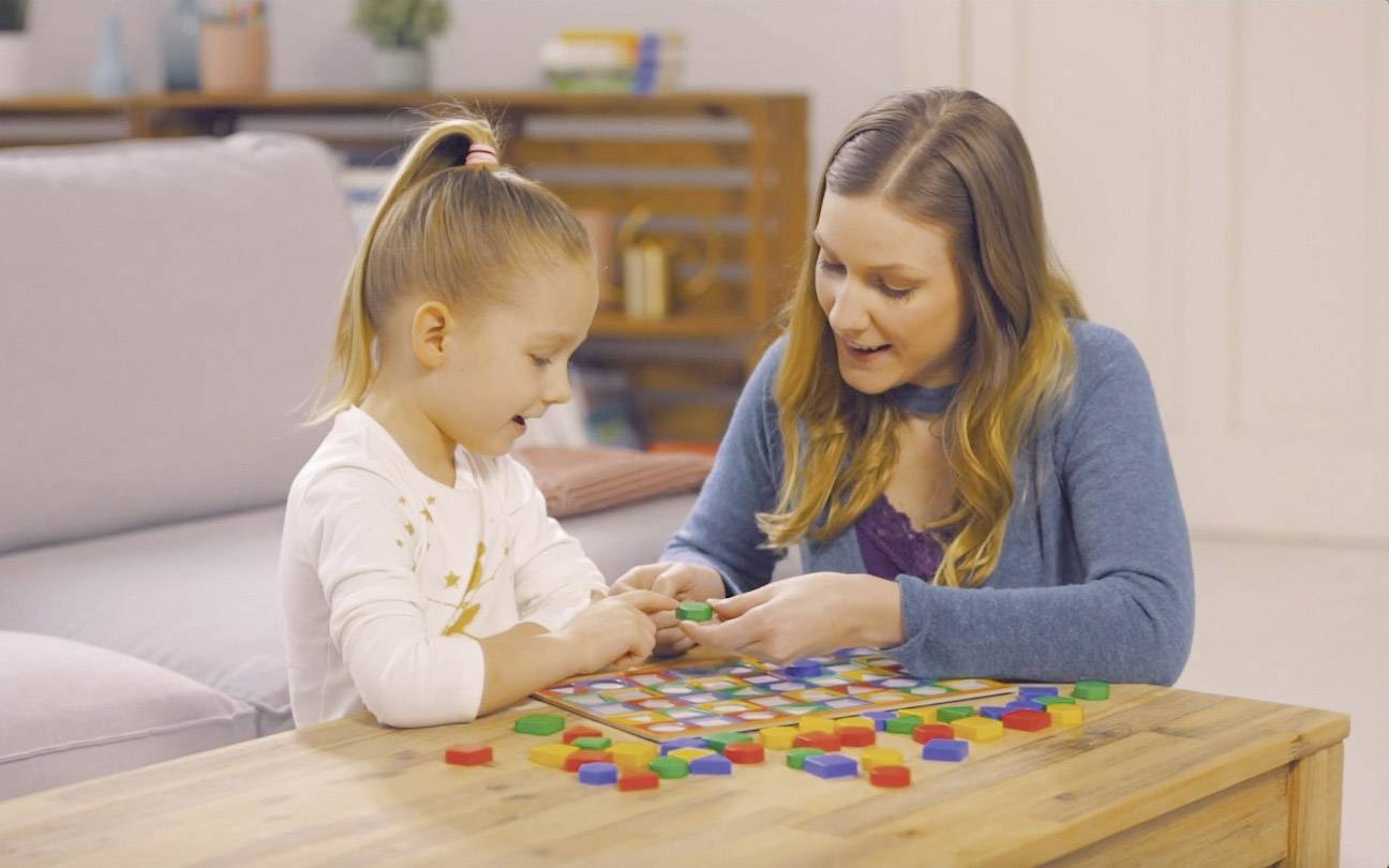 Une personne adulte joue à un jeu de société avec un enfant à une table du salon, tous deux semblant concentrés et joyeux.