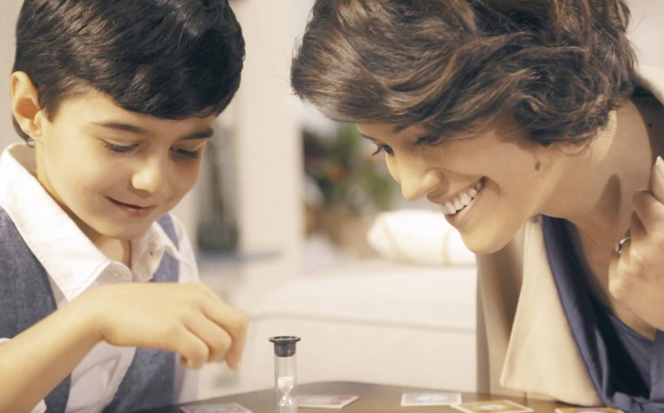 Un garçon et une femme jouent à un jeu de société et rient. Sur la table se trouve un sablier qui pourrait faire partie du jeu.