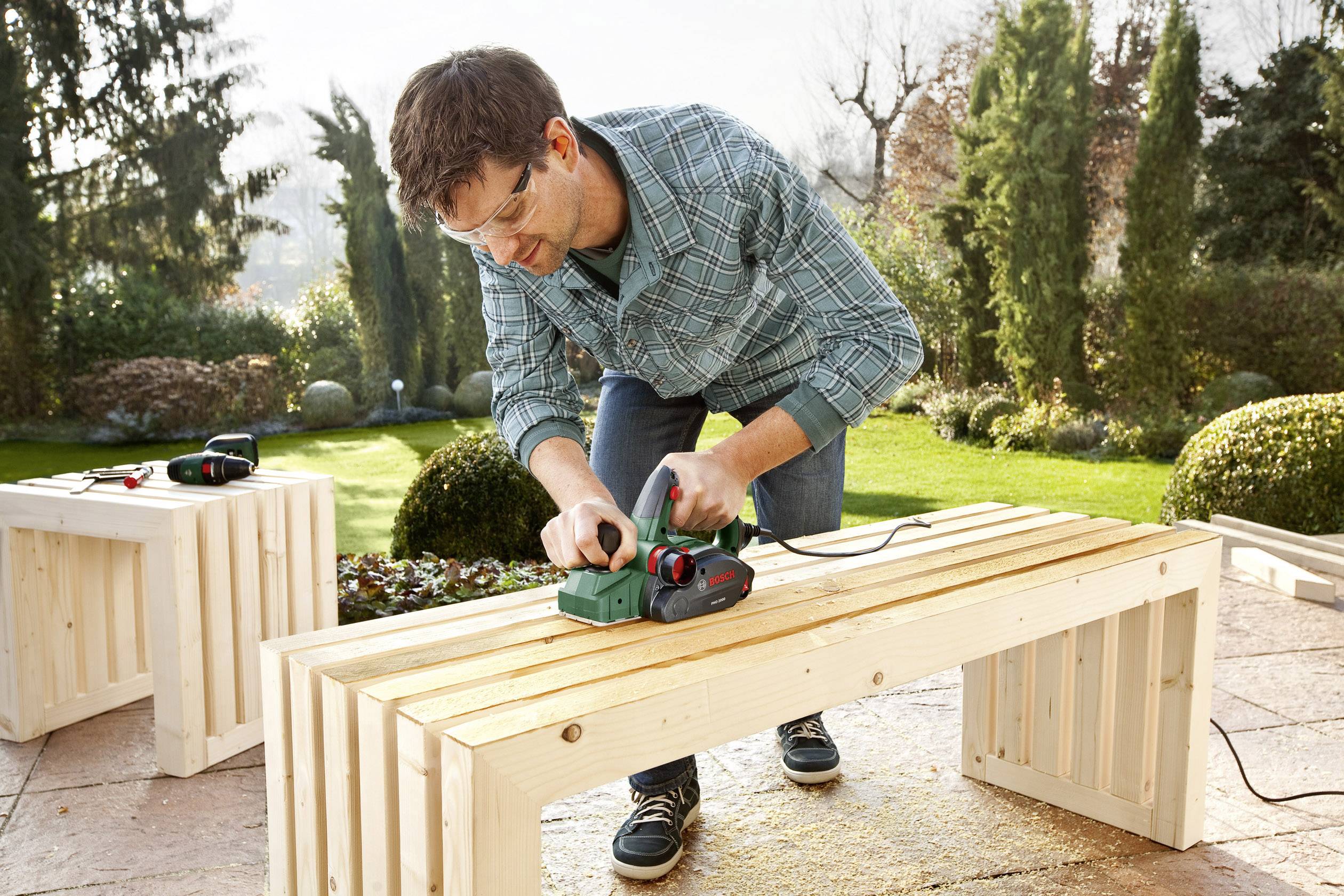 Un homme travaille un banc en bois à l'extérieur avec un outil électrique. Il porte des lunettes de protection.