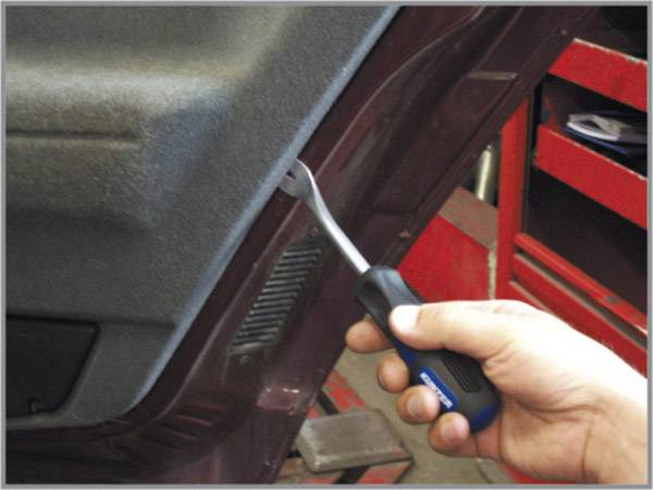 Une main tient un outil pour retirer le revêtement intérieur d'un véhicule. Des étagères d'atelier sont visibles en arrière-plan.