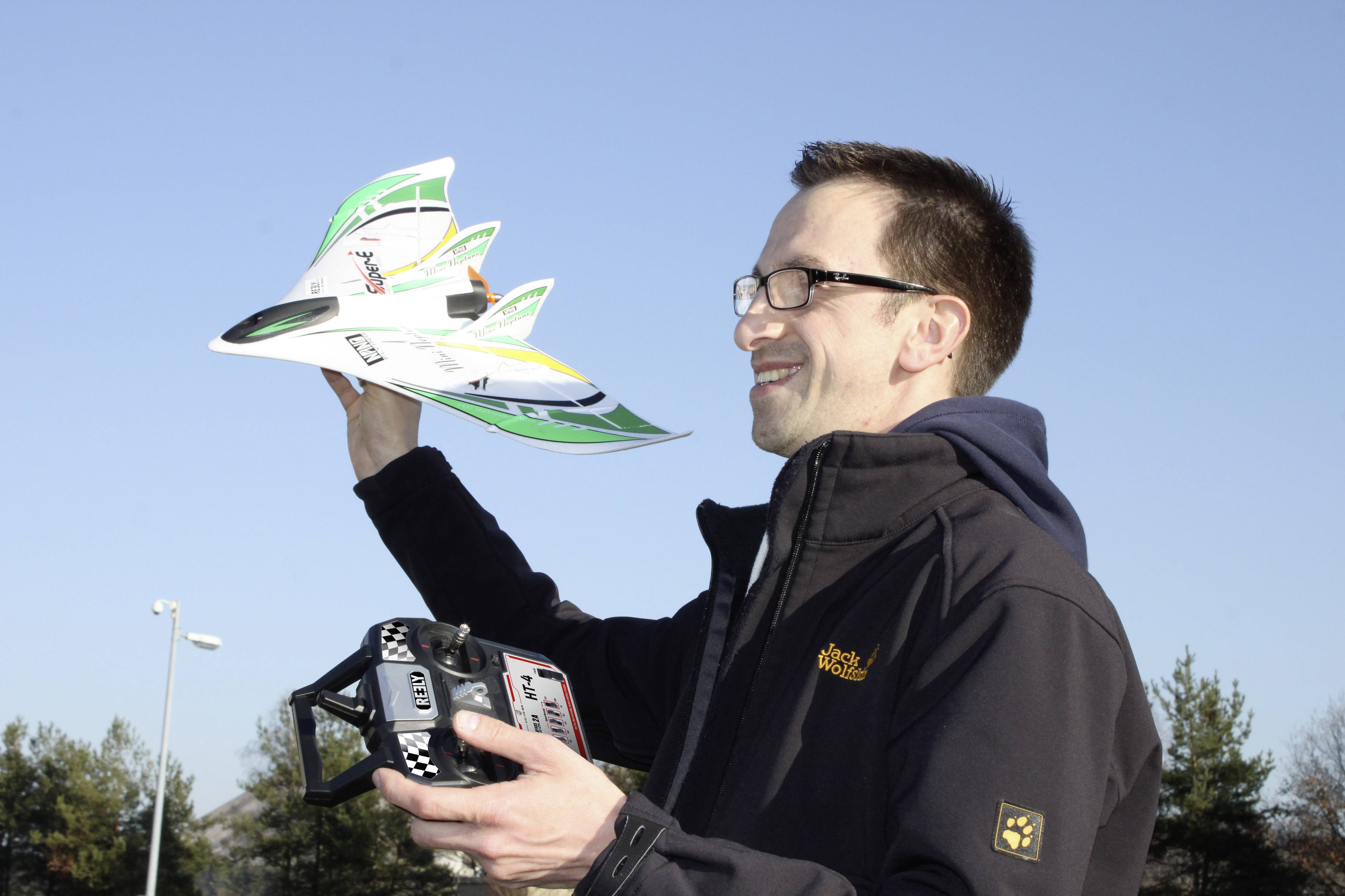 Un homme tient un avion miniature dans sa main droite et une télécommande dans sa main gauche. Des arbres sont visibles en arrière-plan.