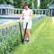 Une femme taille le bord d'un massif de fleurs avec un grand ciseau à haie dans un jardin soigné doté d'une fontaine et de colonnes.