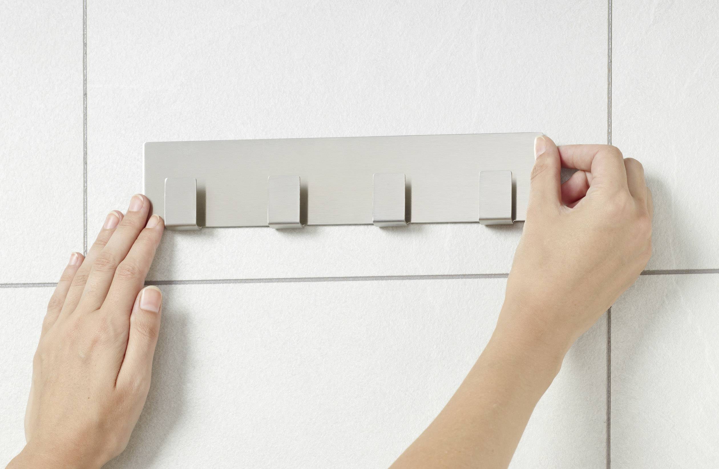 Des mains fixent une barre métallique avec des crochets sur un mur carrelé.