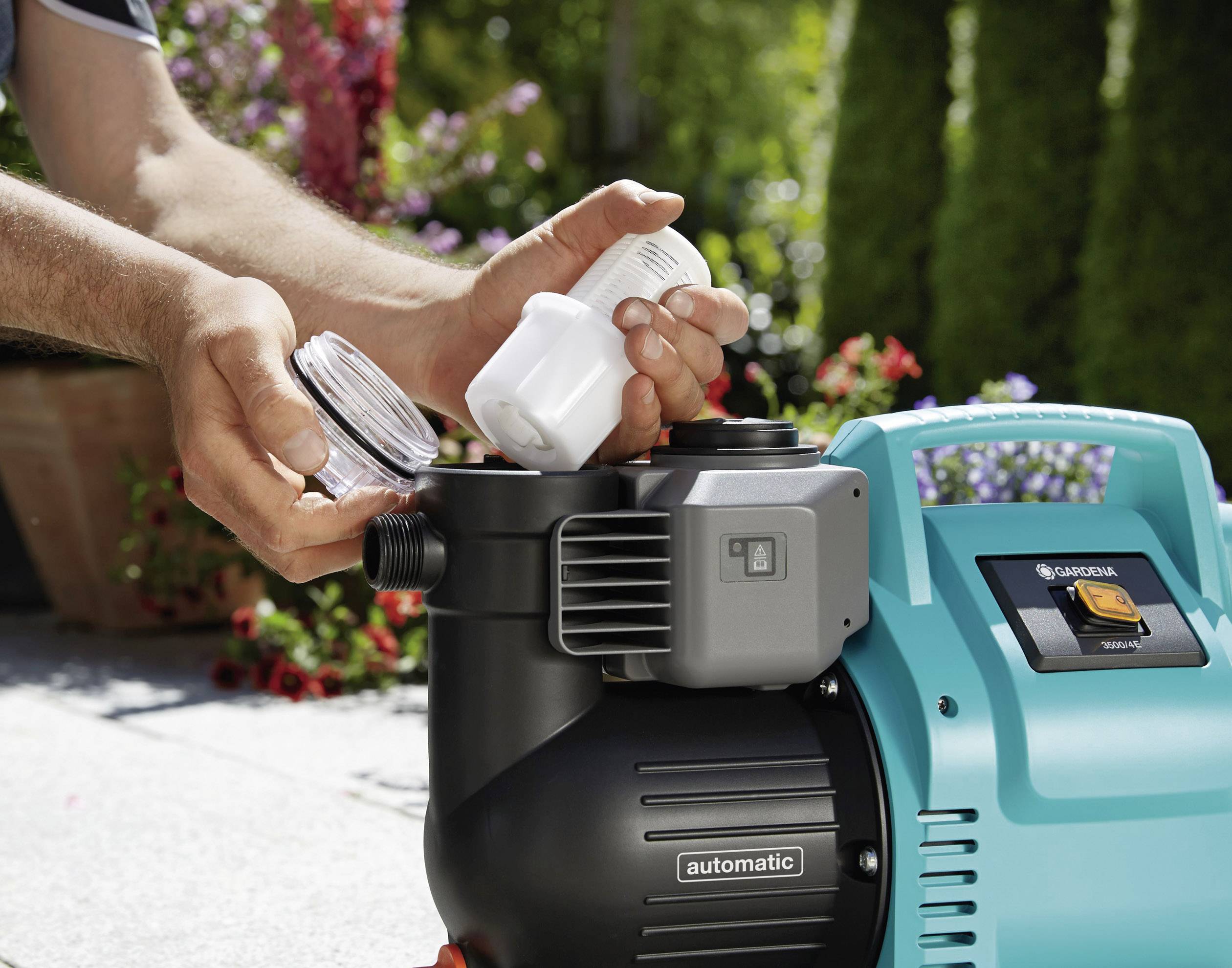 Une personne montre comment installer un filtre à eau dans une pompe de jardin. Des fleurs colorées sont visibles en arrière-plan à gauche de l'image.