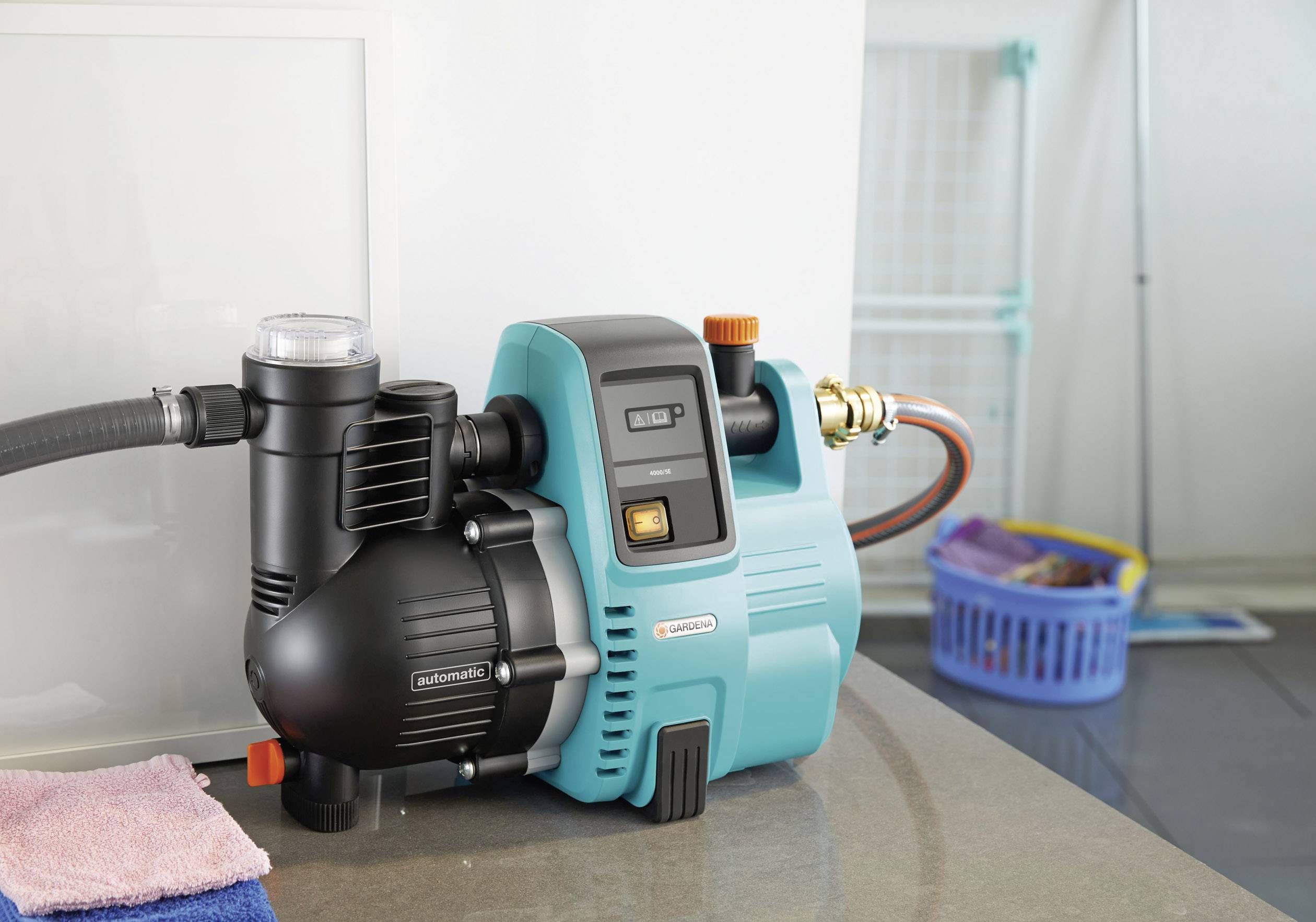 Une pompe à eau électrique est posée sur un plan de travail. Des serviettes sont disposées à côté. À l'arrière-plan, un panier à linge séchoir est visible.