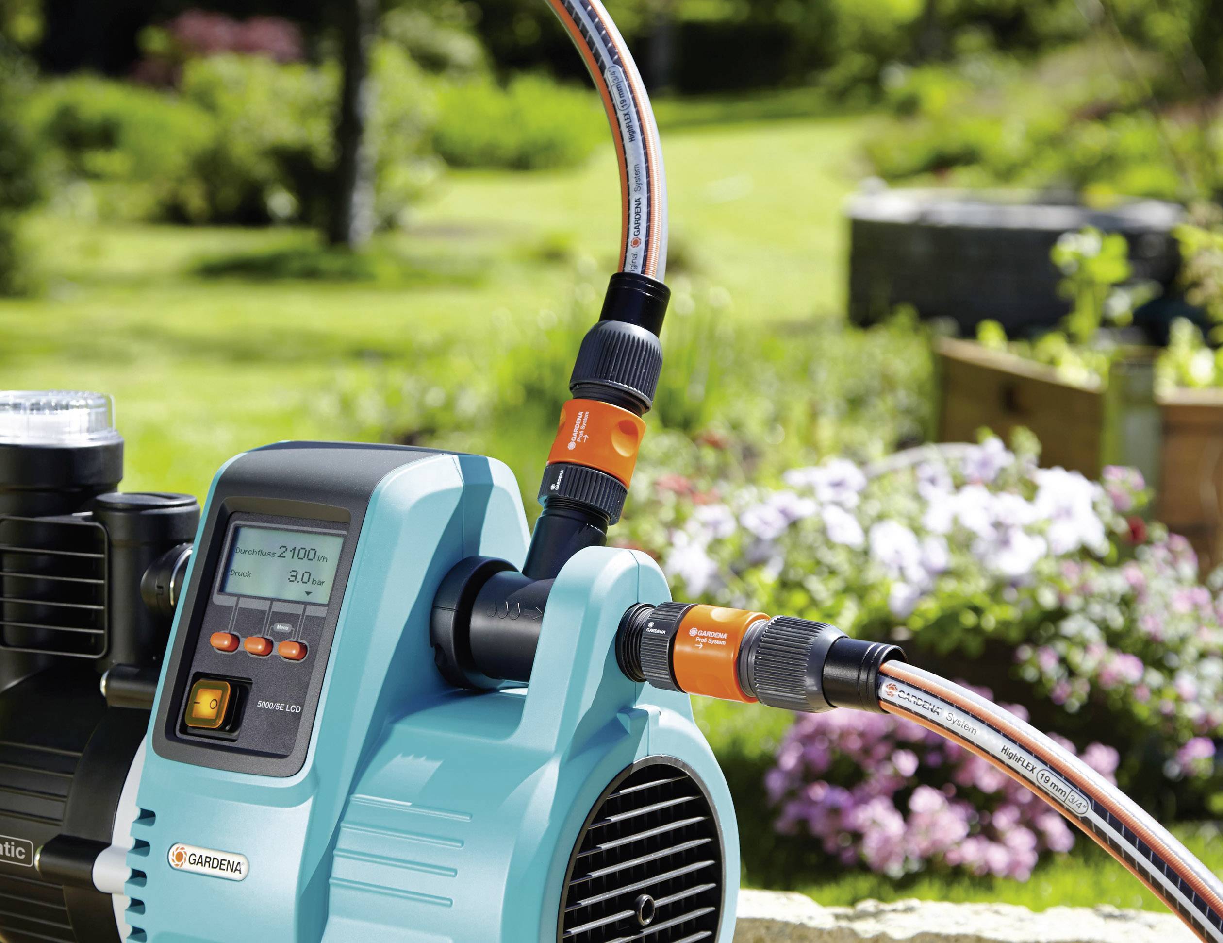 Pompe de jardin Gardena en action, au premier plan avec des raccords de tuyau, un jardin vert et fleuri en arrière-plan.