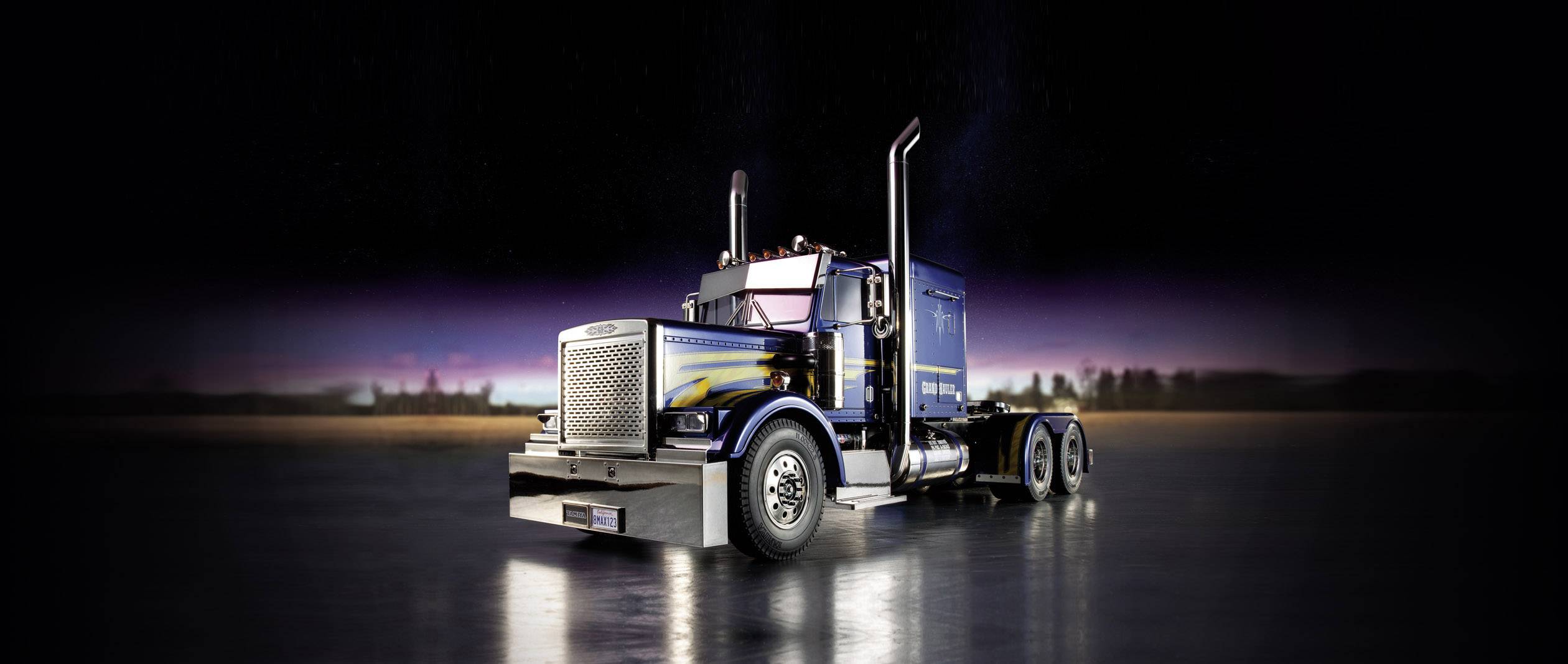 Un camion bleu aux éléments chromés est stationné sur une chaussée mouillée sous un ciel sombre et crépusculaire.