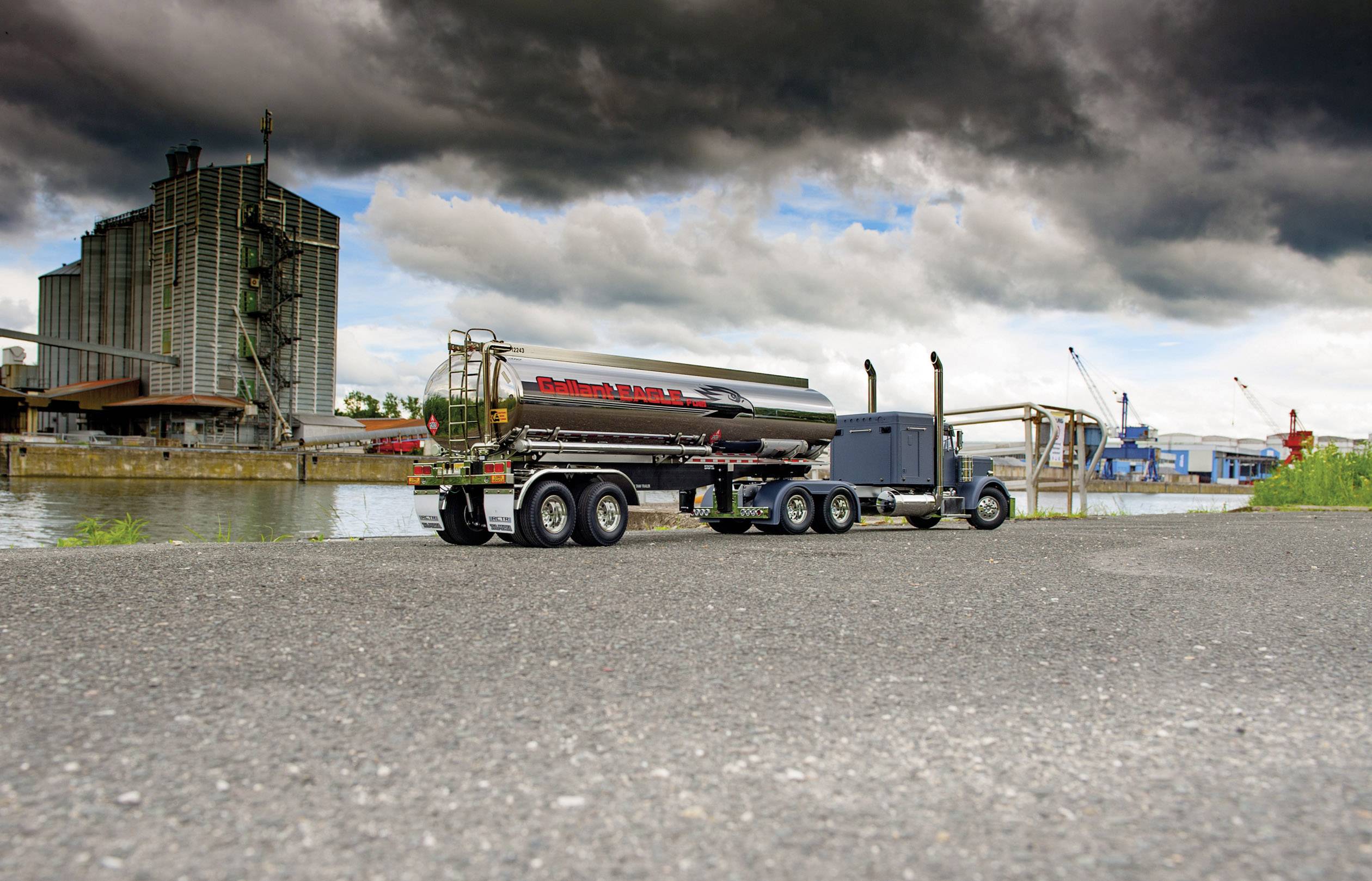 Un camion-citerne modèle avec une remorque-citerne est stationné sur une route en bordure de rivière, devant des nuages gris et des bâtiments industriels en arrière-plan.