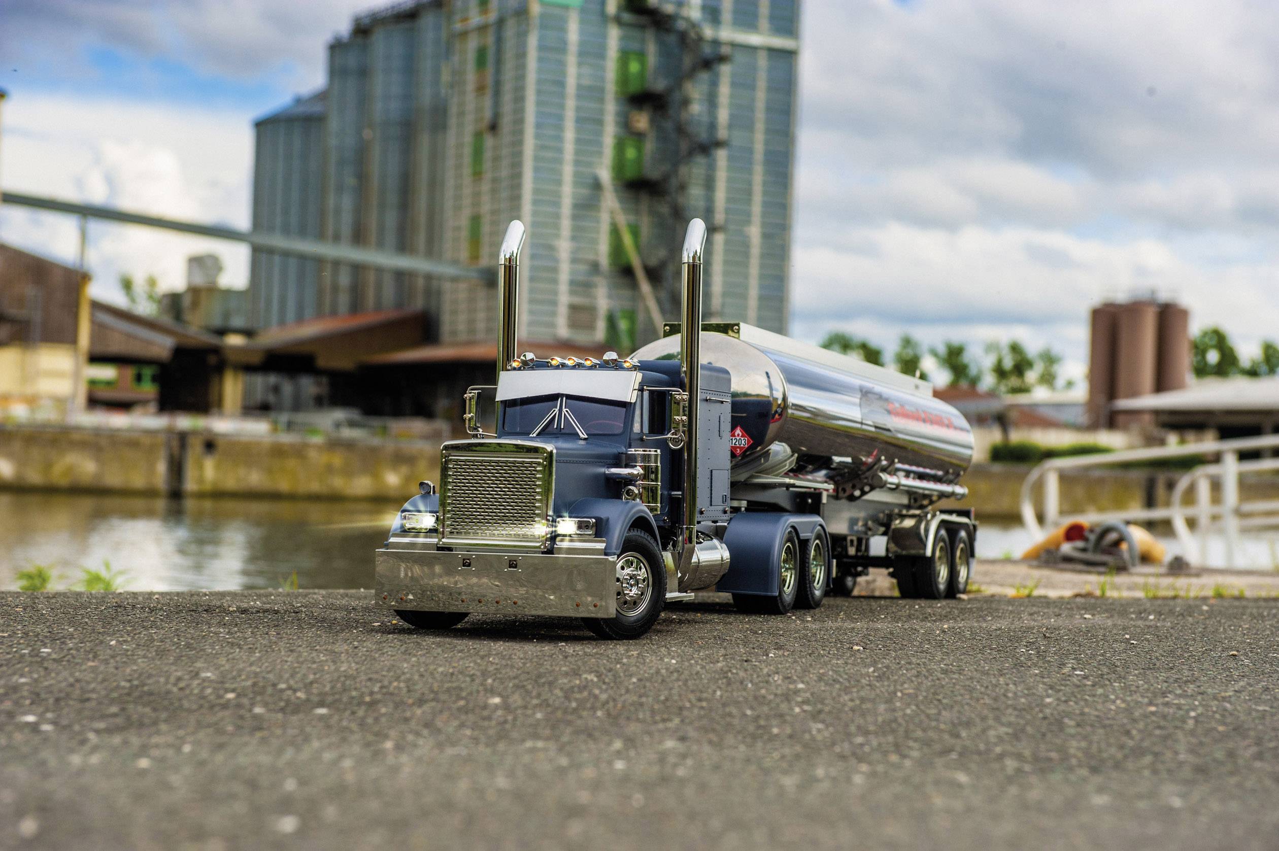 Un camion-citerne miniature est stationné sur une route devant un port, avec des bâtiments industriels en arrière-plan.