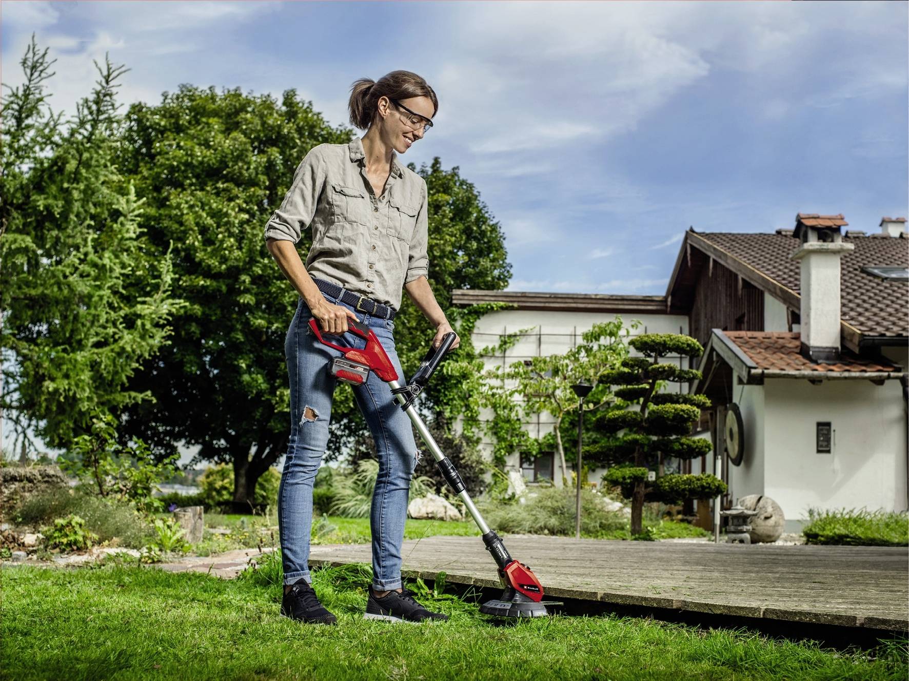 Une femme en tenue décontractée tond la pelouse à côté d'une maison avec un coupe-bordures électrique. Des arbres sont visibles en arrière-plan.