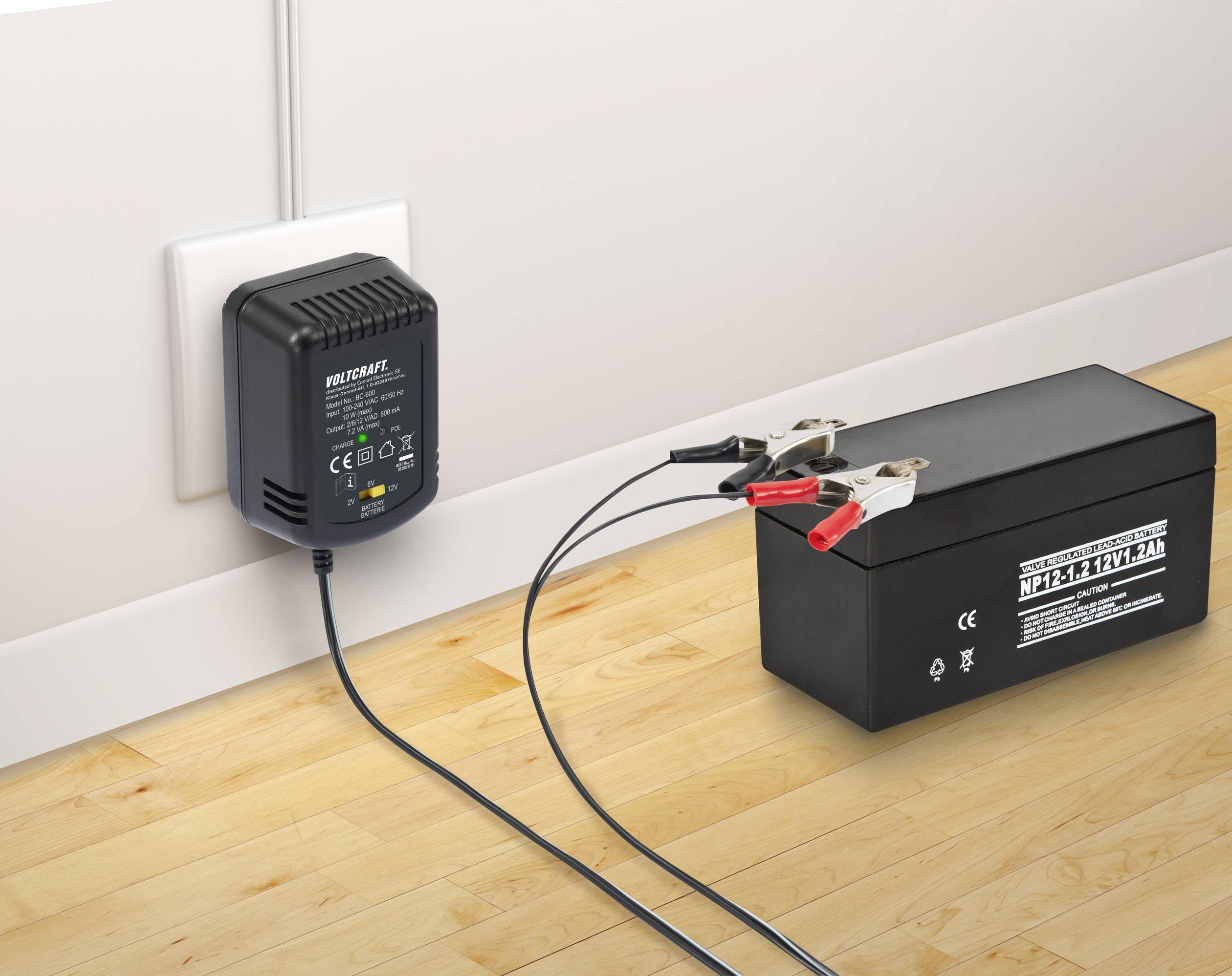 Un chargeur de batterie noir est branché à une prise électrique et charge une batterie de 12V noire à l'aide de pinces sur un sol en bois.