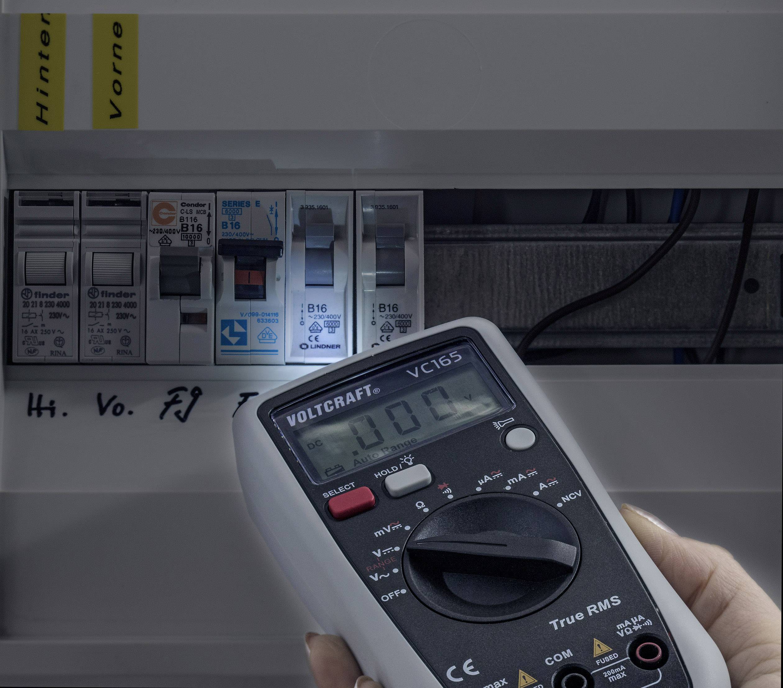 Un appareil de mesure portable mesure 230V dans un tableau électrique ouvert avec plusieurs disjoncteurs, des câblages étant visibles en arrière-plan.