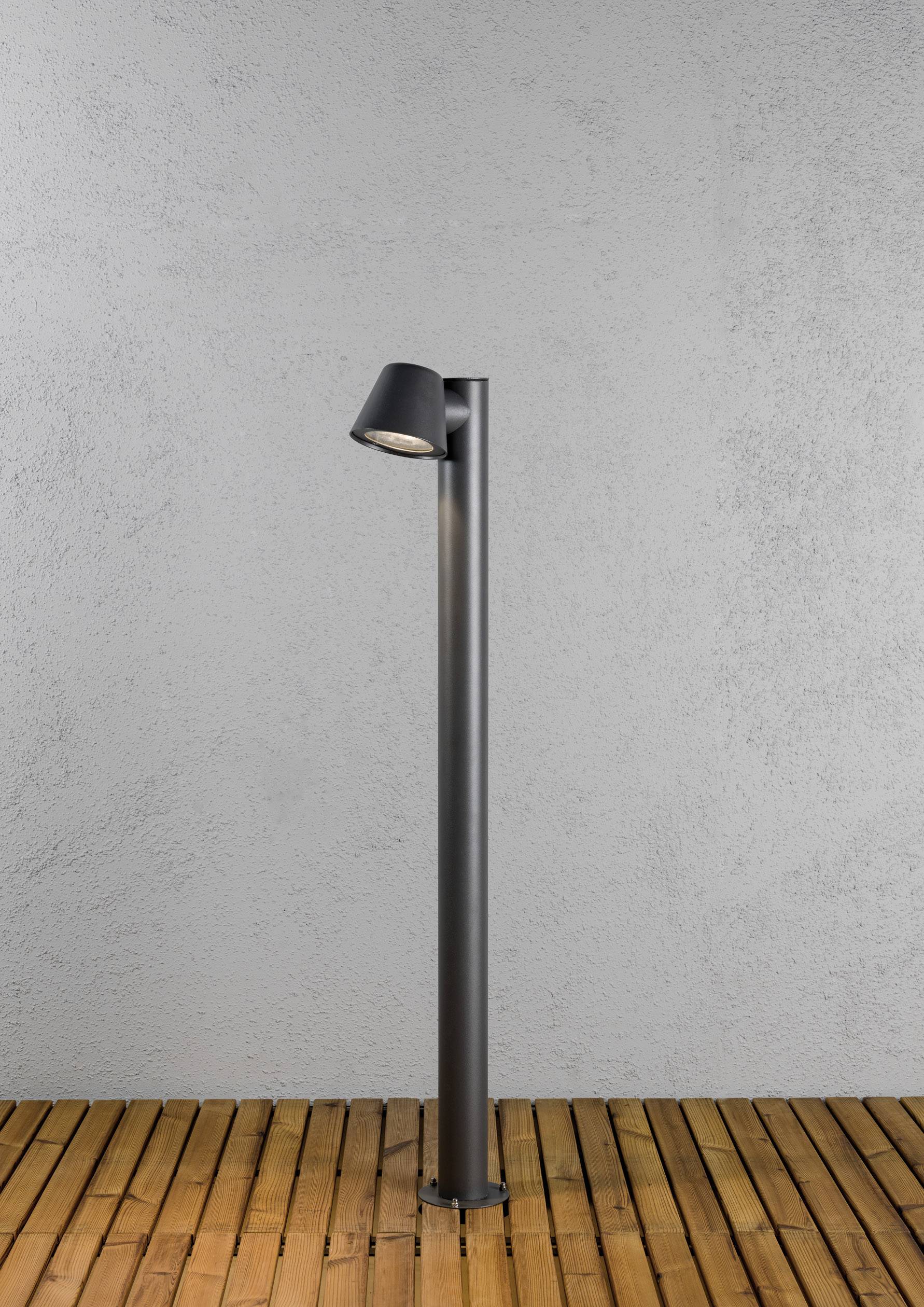 Un luminaire extérieur moderne noir sur une terrasse en bois devant un mur gris. Le luminaire possède un socle cylindrique simple et épuré.