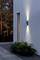 Applique murale extérieure illuminant un trottoir contre une façade blanche moderne. Des arbres sont visibles en arrière-plan au crépuscule.