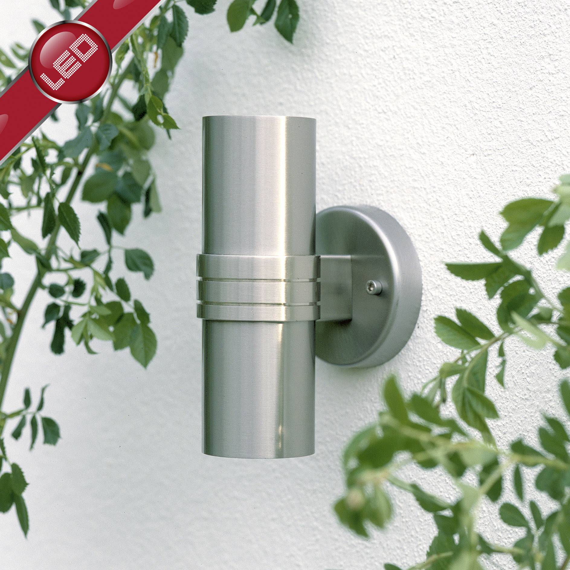 Applique murale moderne en acier inoxydable brossé, de forme cylindrique, montée sur un mur blanc, entourée de plantes vertes.