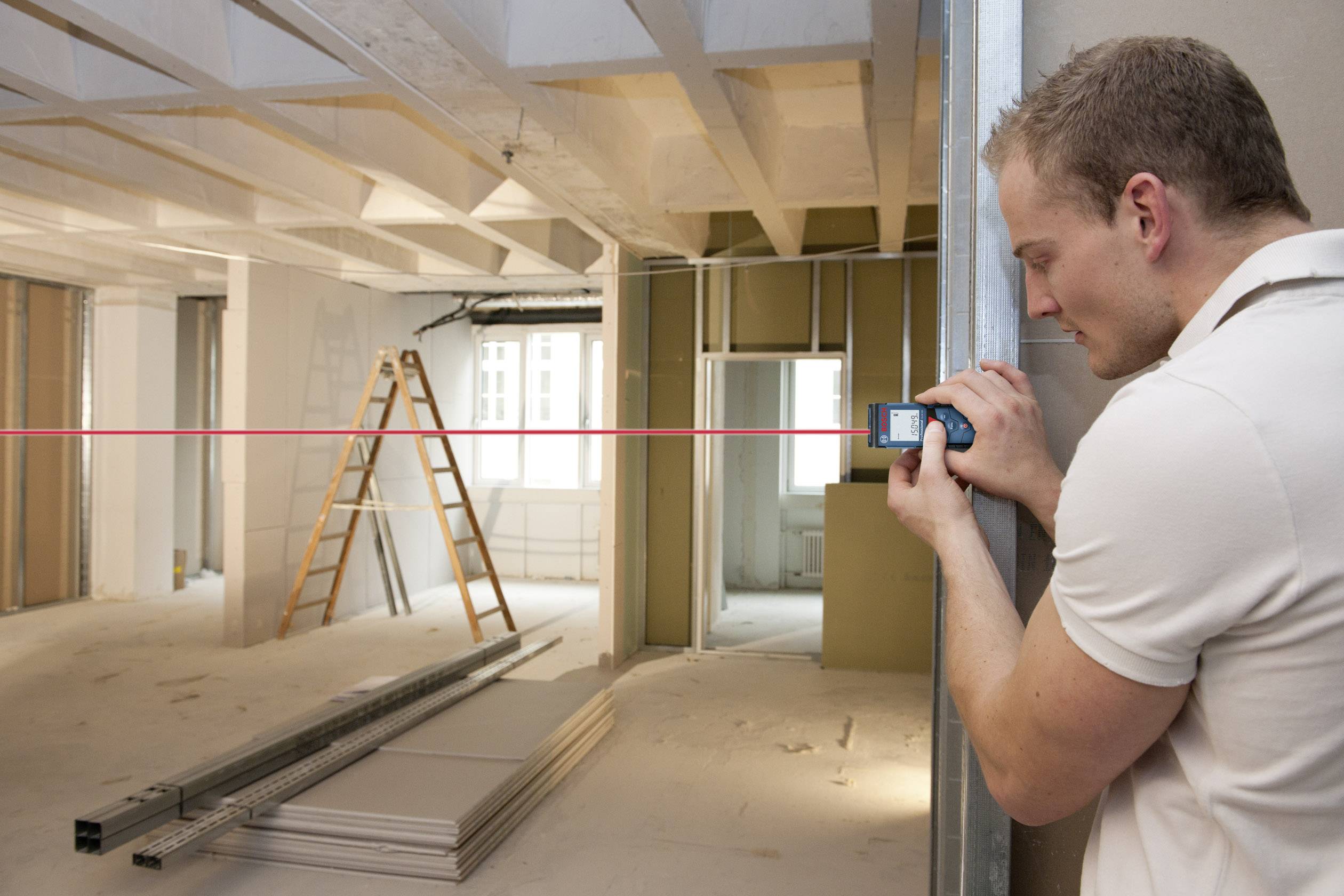 Un homme portant un t-shirt blanc mesure une pièce avec un télémètre. La pièce est en travaux, avec des échelles et des matériaux de construction.