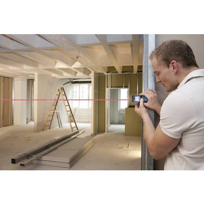 Un homme portant un t-shirt blanc mesure une pièce avec un télémètre. La pièce est en travaux, avec des échelles et des matériaux de construction.