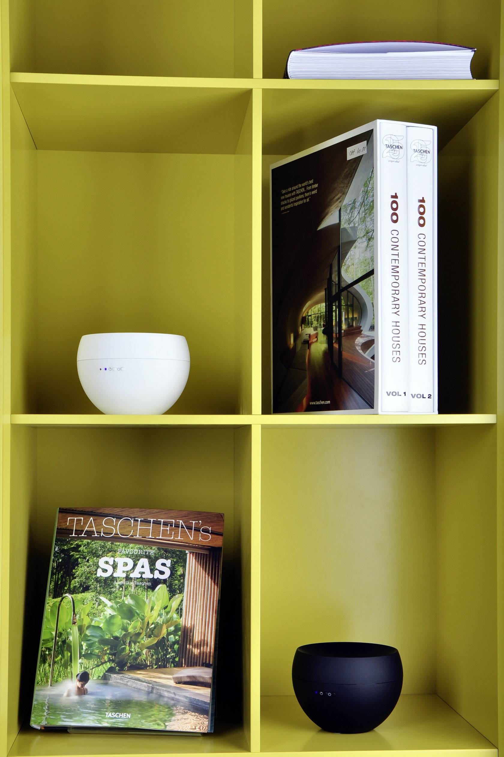 Une étagère jaune avec deux livres '100 Contemporary Houses', un magazine 'TASCHEN's SPAS' et deux enceintes rondes.