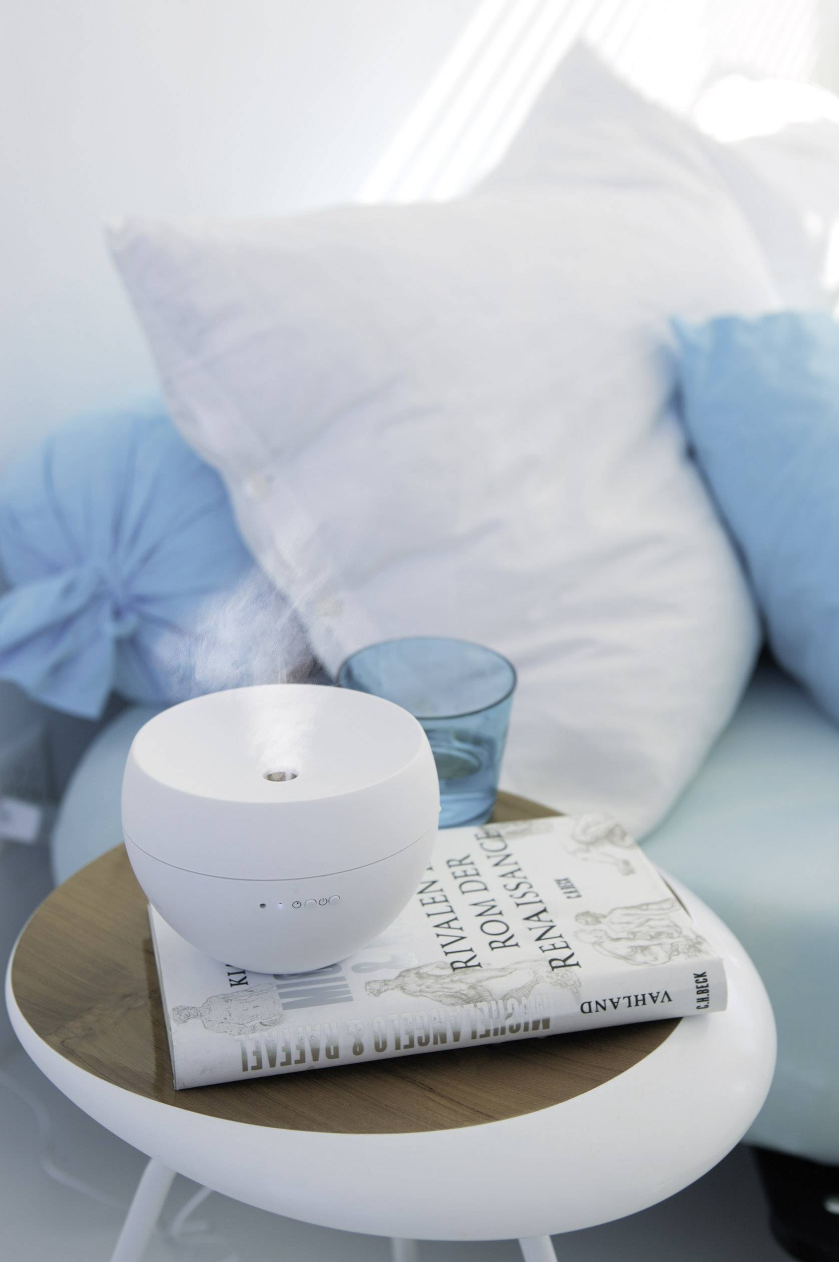 Un humidificateur est posé sur une petite table à côté d'un livre intitulé 'Rallye des Rivaux Aventure'. Un canapé avec des coussins se trouve en arrière-plan.
