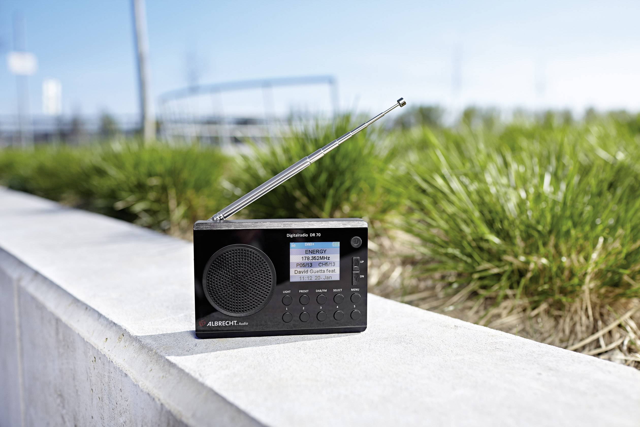 Une radio portable avec une antenne télescopique est posée sur un mur en béton. En arrière-plan, on aperçoit de l'herbe verte floue.
