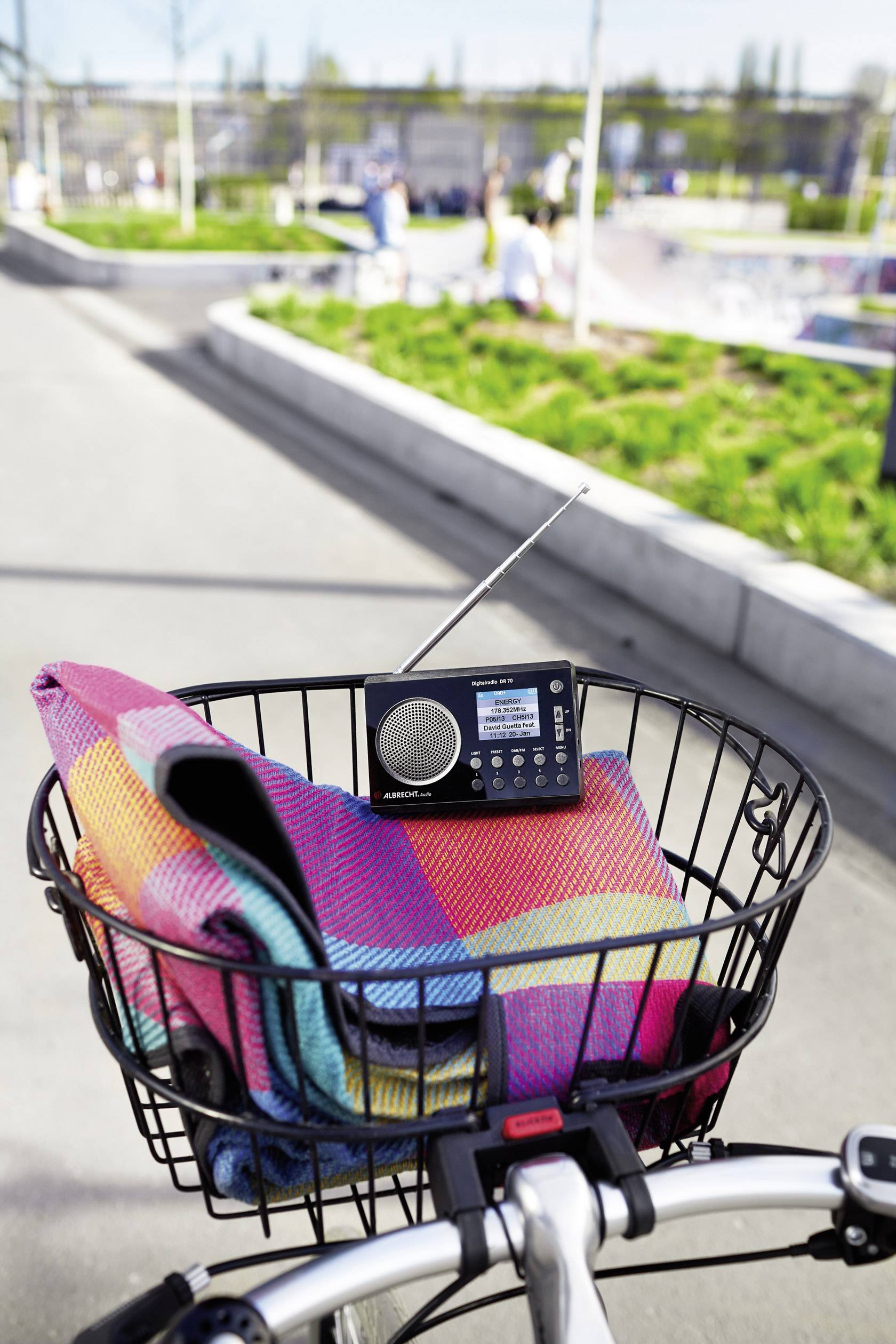 Un vélo avec un panier noir contient une petite radio et une couverture colorée. En arrière-plan, on aperçoit un parc avec des espaces verts.