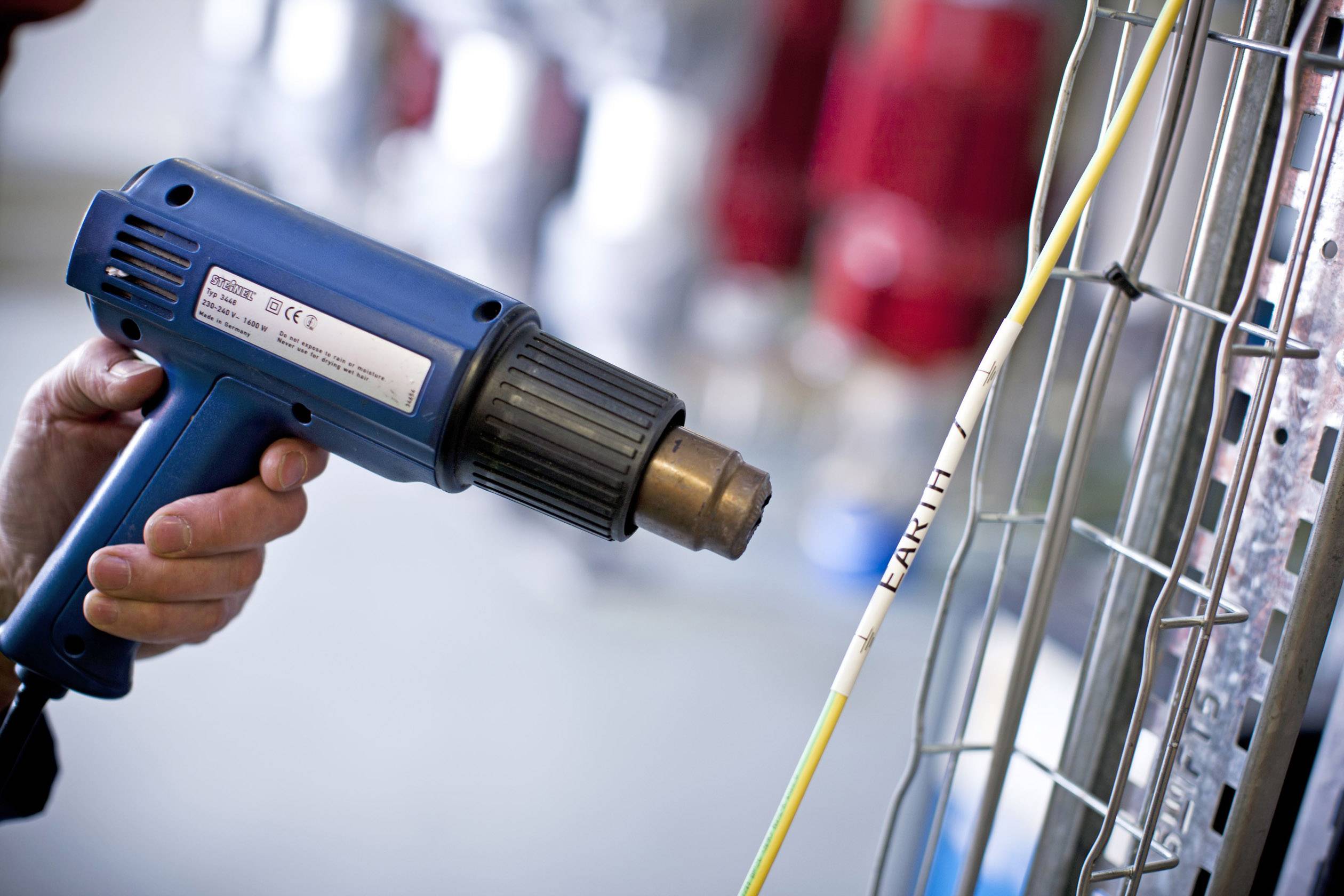 Une personne utilise un pistolet à air chaud bleu pour sceller des gaines thermorétractables sur des câbles. Des objets flous sont visibles en arrière-plan.