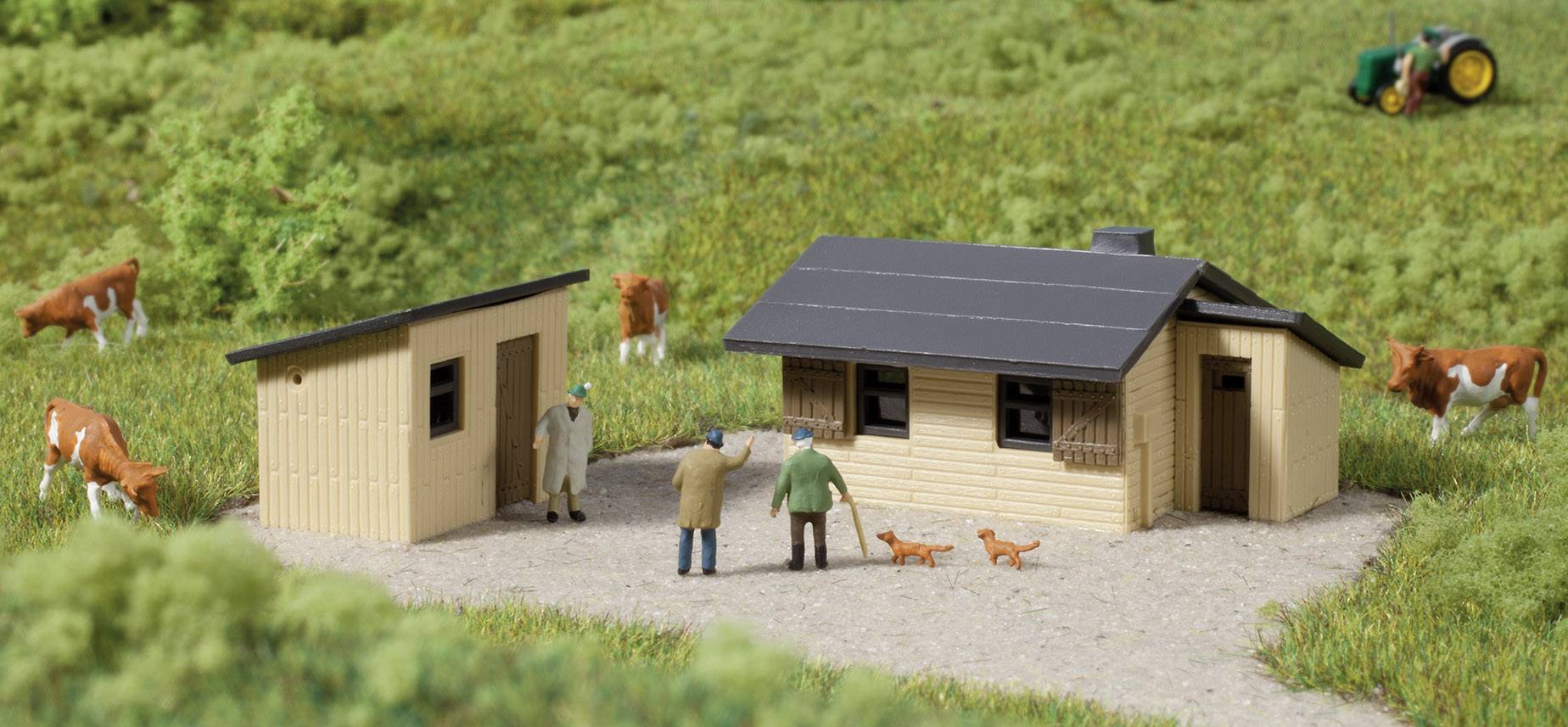 Petit diorama d'une ferme miniature avec deux bâtiments, des personnages, deux chiens et des vaches sur un champ vert. Un tracteur est visible en arrière-plan.