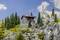 Une cabane de montagne dans un paysage alpin avec des arbres et des rochers environnants, une croix sommitale se dresse sur une montagne à l'arrière-plan sous un ciel bleu.