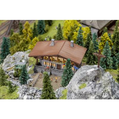 Une petite maison de montagne miniature est entourée d'arbres. Des personnes marchent le long d'un chemin. Des voies de chemin de fer miniatures sont visibles en arrière-plan.