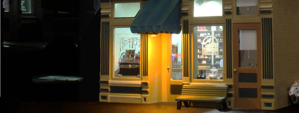 Petit magasin avec une vitrine éclairée, un auvent bleu et un banc en bois devant. Quelques produits sont visibles dans la fenêtre.