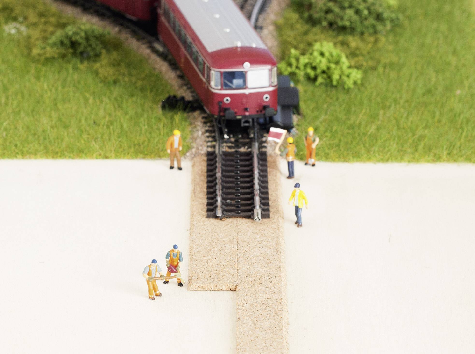 Un train miniature circule sur des rails à travers un paysage. Des figurines miniatures travaillent le long de la voie près d'un passage à niveau.